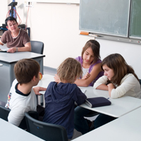 Vier Schülerinnen und Schüler sitzen sich gegenüber und unterhalten sich während einer Gruppenarbeit. Im Hintergrund sitzt die Lehrerin am Schreibtisch neben der Tafel.