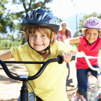 Zwei Kinder radeln lächelnd. Das Kind im Vordergrund fährt ein schwarzes Fahrrad, trägt ein gelbes T-Shirt und einen blauen Helm. Das Kind im Hintergrund fährt ein weißes Fahrrad, trägt ein gelbes T-Shirt und einen fliederfarbenen Helm.