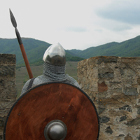 Mit dem Rücken zum Fotografen schaut ein Ritter mit Schild und Speer von den Zinnen einer Burg.