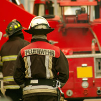 Vor einem Feuerwehrauto stehen zwei Feuerwehrmänner mit dem Rücken zum Fotografen.