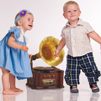 Zwei Kleinkinder bewegen sich zur Musik aus einem alten Grammophon, welches auf dem Boden steht.